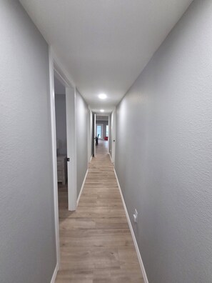 A long, narrow hallway with light gray walls and wooden flooring, leading to a room with a red chair and a white bed.