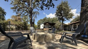 Un área de estar al aire libre con sillas Adirondack, un hogar de piedra y vista a un edificio de fondo.