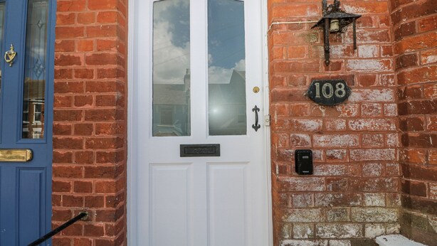 A white door with a glass panel, a blue door to the left, and a brick wall with a house number sign. - Thistle Villa (Hastings)
