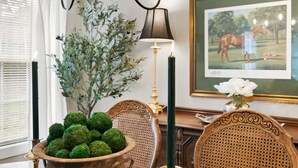 A room with a wooden chair, a table with a bowl of green balls, a lamp, and a framed picture of a horse.