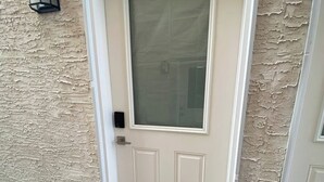 A beige textured wall with a white door featuring a frosted glass window and a small rectangular panel. A wooden bench is placed at the door's entrance. - Guest Suite (Winnipeg)