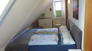 A small, cozy room with a sloped ceiling, a bed with multiple pillows, a small table, a cabinet, a window with blinds, and a painting on the wall. - NEW! Vacation apartment Sonnenschein OG (Neuschoo)