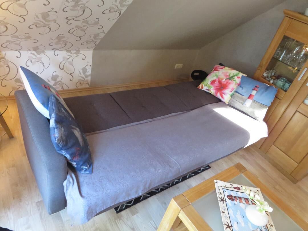 Ein kleines Zimmer mit einem Einzelbett, einer grauen Tagesdecke, einem floralen Kopfkissen und einem Holzkasten mit Glastüren.