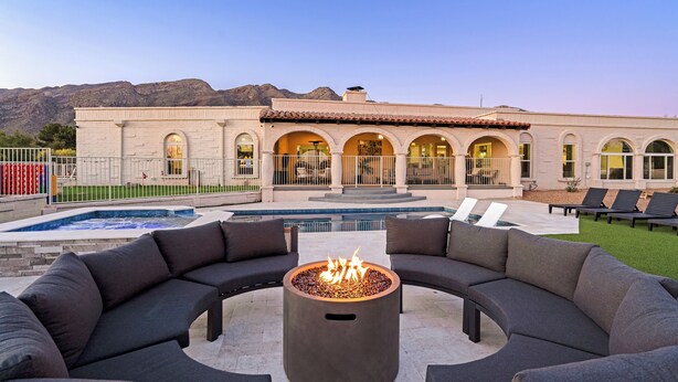 A pool area with a fire pit, outdoor seating, and a building with arched windows. - Luxury 5BR Foothills Home Heated Pool & Spa, Gym, Mountain Views Sleeps 16 (Tucson)