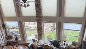 Ein geräumiges Wohnzimmer mit einer Ledersofa, einem Holztisch und einem Fernseher. Mehrere Fenster sorgen für natürliches Licht und Blick ins Freie.
