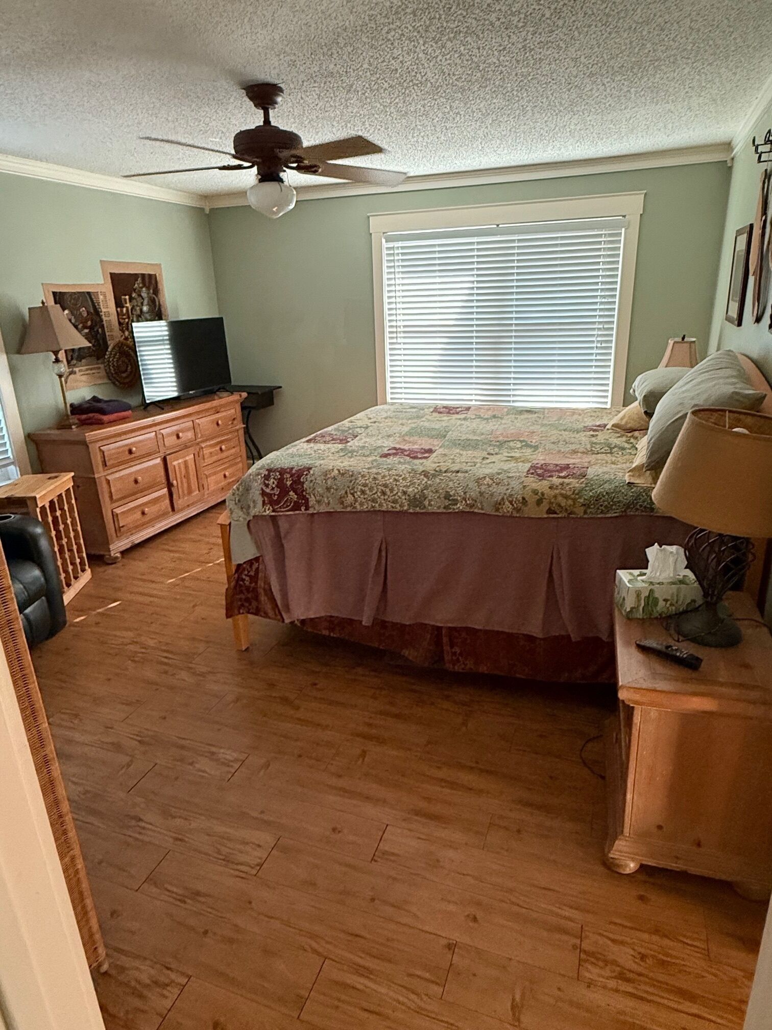 A bedroom with a bed, a ceiling fan, a window with blinds, a dresser, a television, and a lamp.