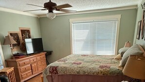 Ein Schlafzimmer mit einem Bett, einem Deckenventilator, einem Fenster mit Jalousien, einem Schminktisch, einem Fernseher und einer Lampe.