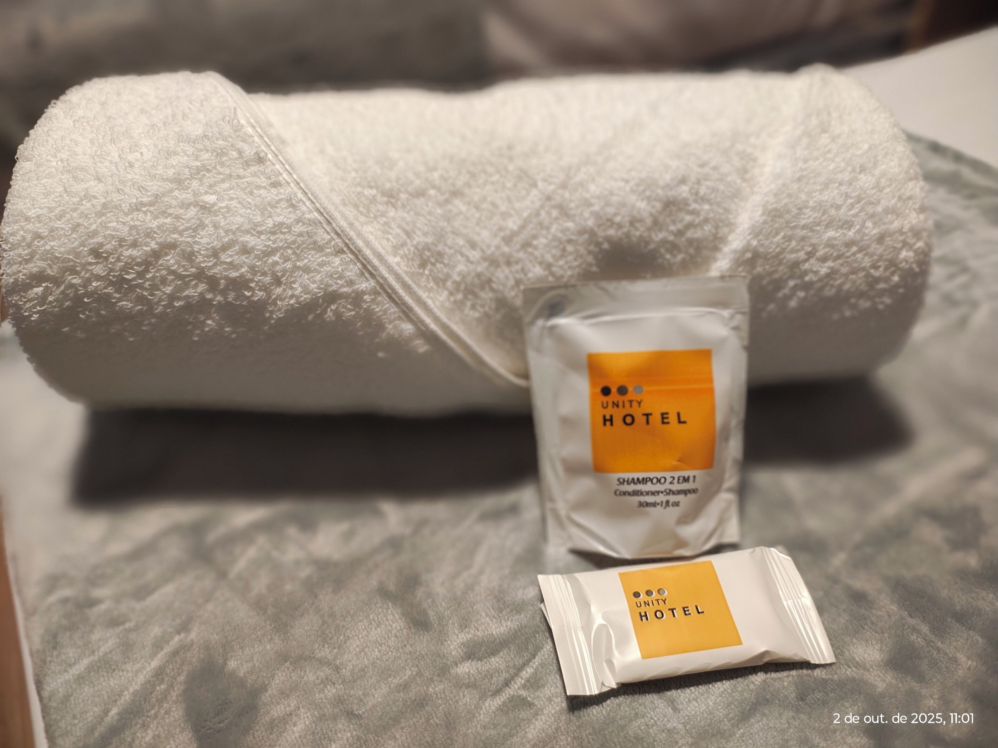A neatly folded white towel and a small bottle of shampoo with a Unity Hotel label on a textured bedspread.