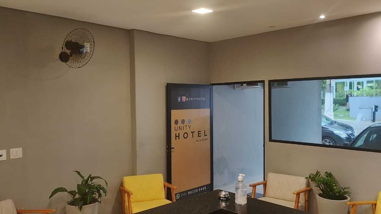 Un salón de conferencias con una mesa, sillas, un ventilador, un cartel y una ventana que ofrece vistas de un edificio y un coche.