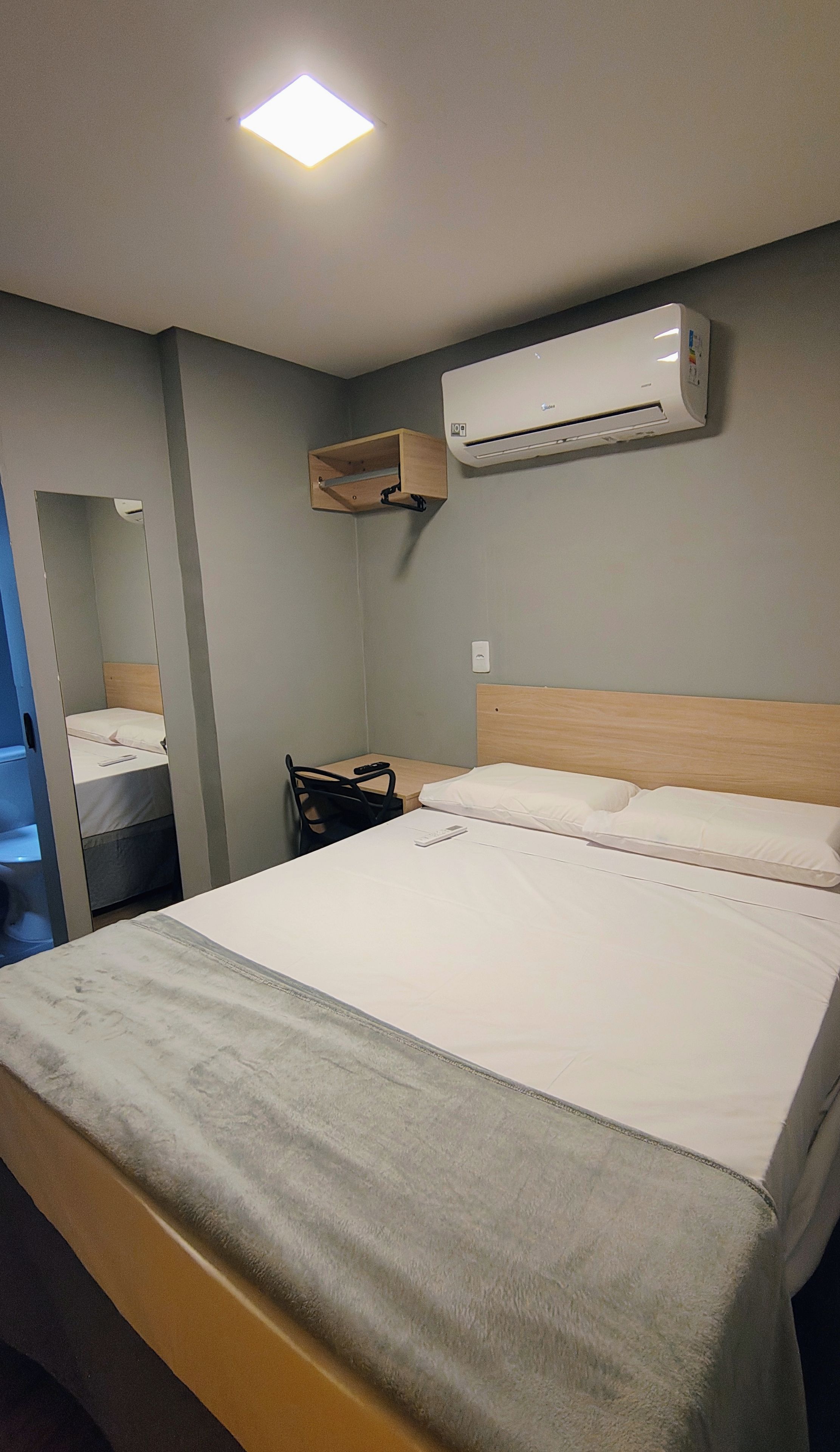 A hotel room with a bed, a wooden headboard, an air conditioning unit, a small wooden shelf, a mirror, and a nightstand.
