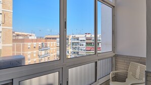 A balcony with a view of urban buildings, a white chair, and a window with blinds.