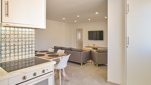 A modern kitchen with a stove, oven, and white cabinets. There is a dining area with a table and chairs, and a gray sofa. A TV is mounted on the wall.