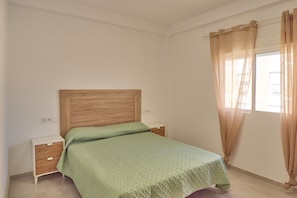 Quarto com cabeceira de madeira, colcha verde, mesa de cabeceira e janela com cortinas bege.