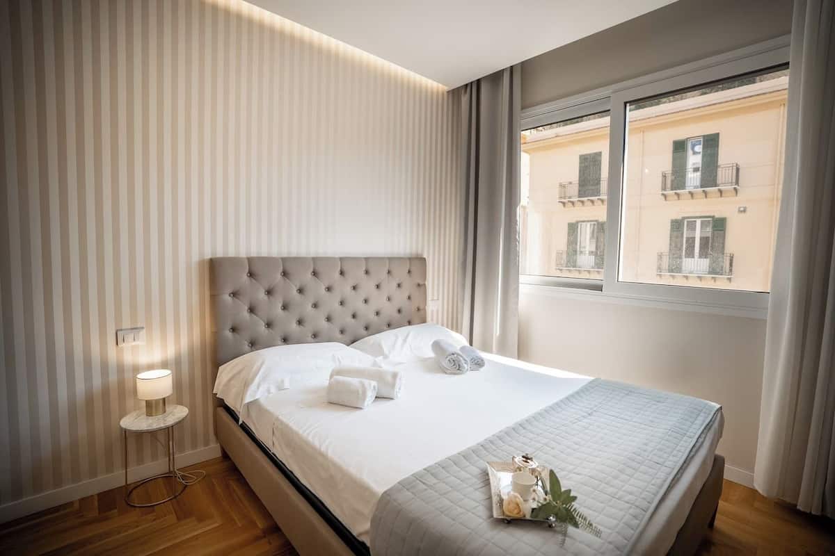 A neatly made bed with pillows and towels, a bedside lamp, and a tray with a cup and decorative items on the windowsill.