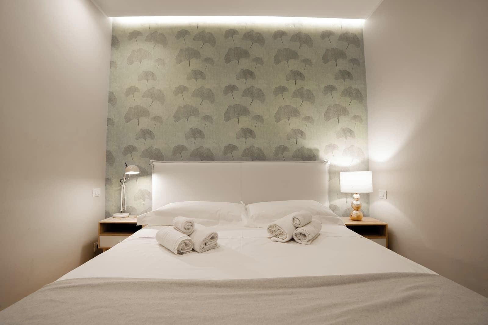 Una cama bien hecha con dos toallas enrolladas, un cabecero blanco y una pared con un diseño de hojas.