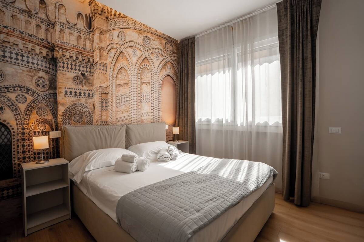 A hotel room with a large bed, a nightstand, a wall tapestry, and a window with curtains.