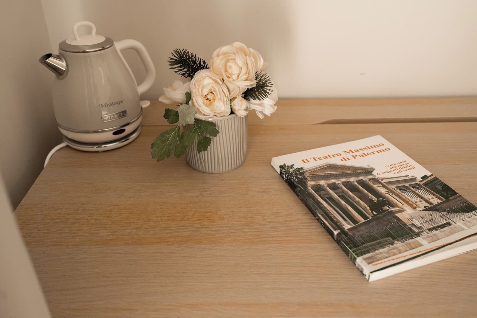 Ein Holztisch mit einer Kanne im Vintage-Stil, einer gestreiften Tasse mit Blumen und einem Buch mit dem Titel 'Il Teatro Massimo di Palermo'.