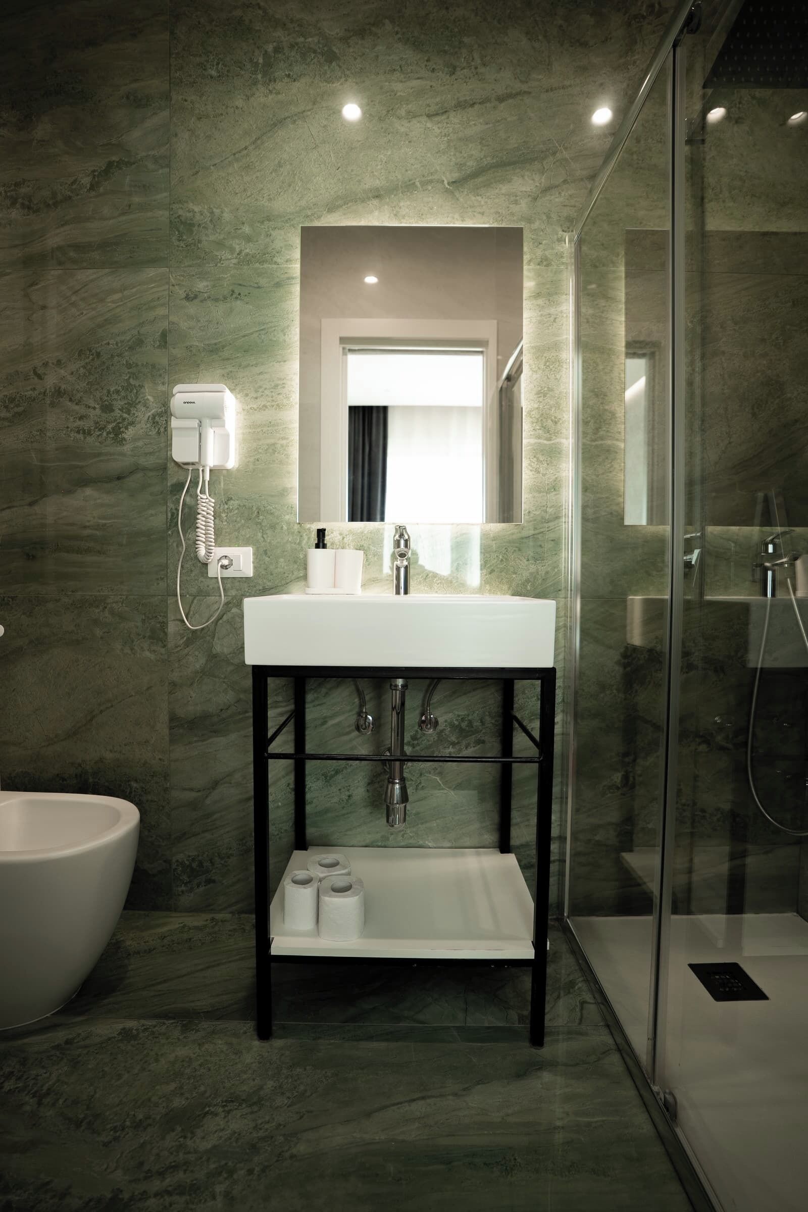 Un baño moderno con un lavamanos blanco, cabina de ducha con mampara de vidrio y pared de mármol.