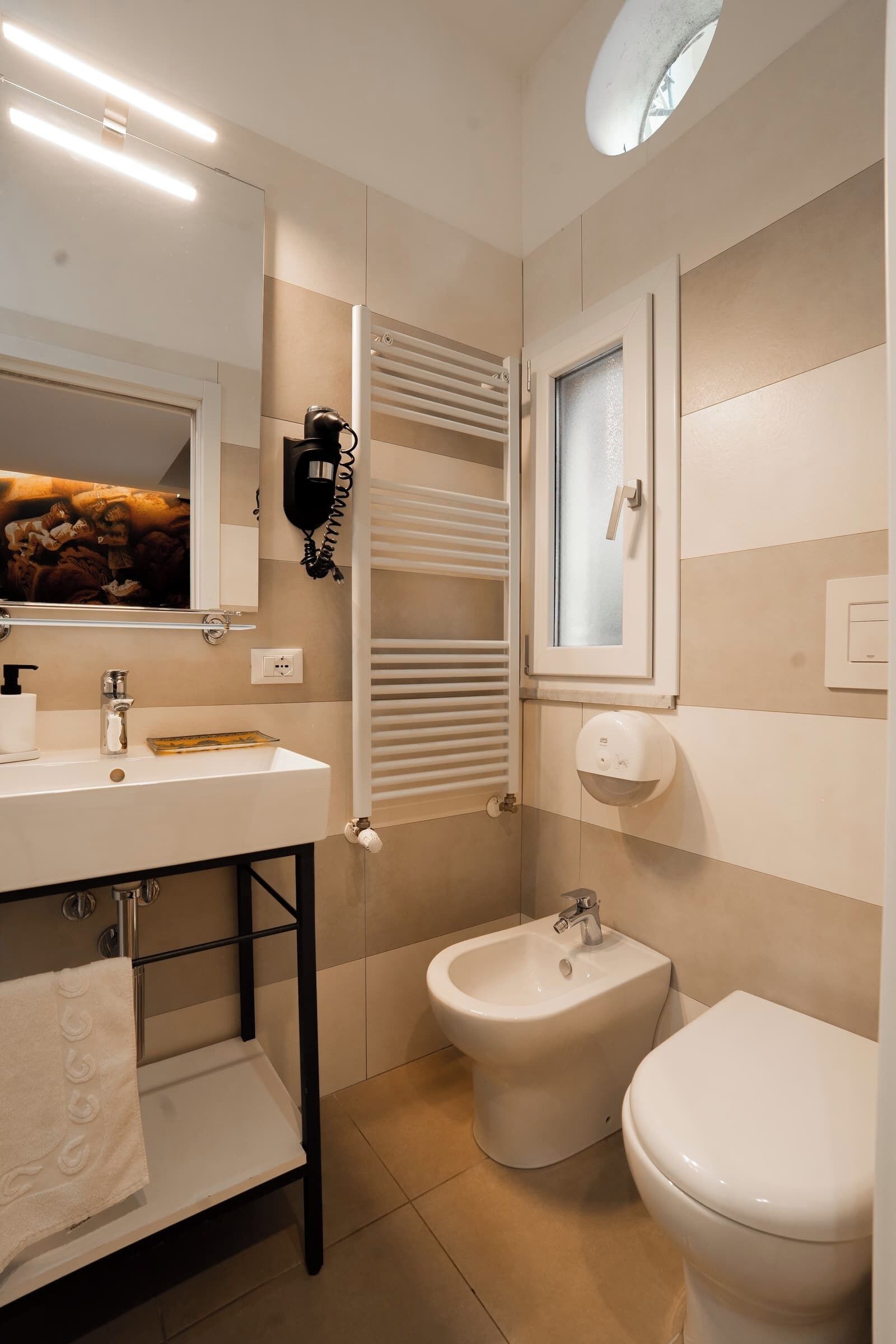 A modern bathroom with a white sink, bidet, and toilet, a towel rack, and a window with blinds.