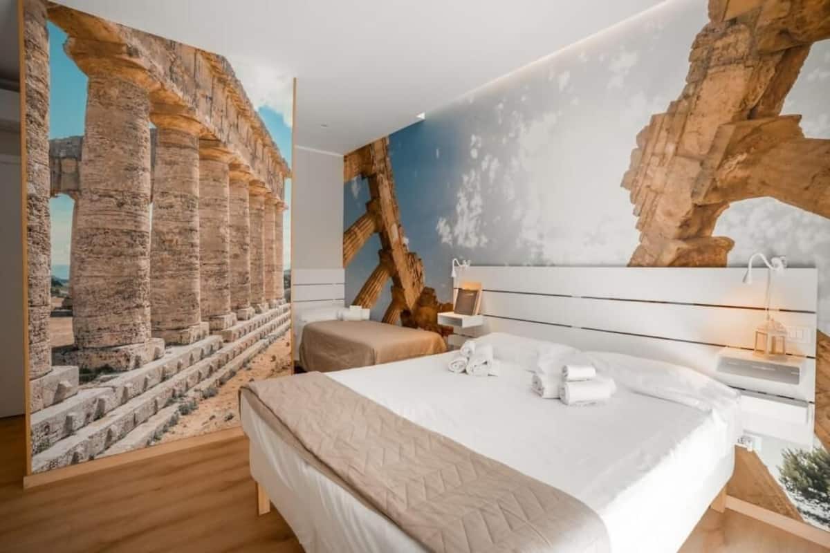 A modern bedroom with a large bed, a nightstand, and a wall mural of ancient ruins.