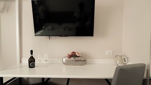 Habitació d'hotel moderna amb televisió plana penjada a la paret, taula blanca amb una ampolla de vi, dues copes i un ram de flors decoratiu.
