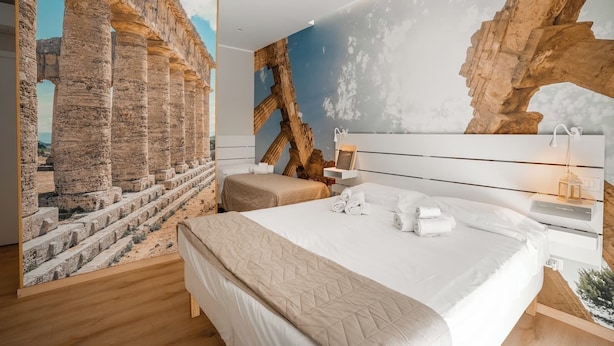 Habitació d'hotel amb un mural gran de ruïnes antigues, un llit amb roba de llit blanca, una tauleta de nit amb una làmpada i un terra de fusta.