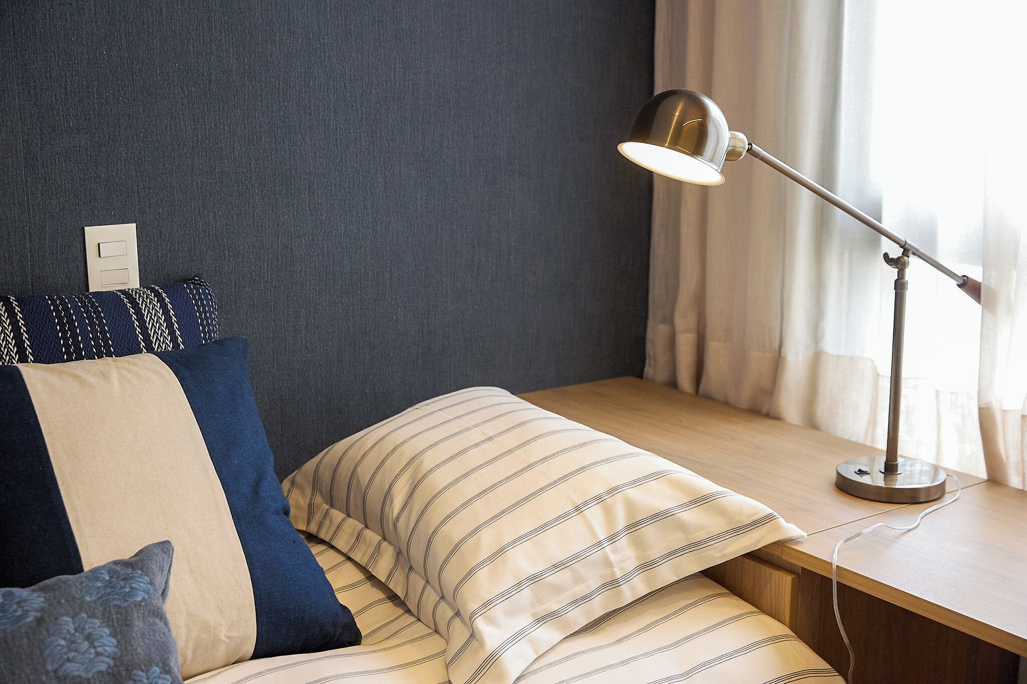 Una cama bien hecha con ropa de cama a rayas, una lámpara de noche y un enchufe en la pared.