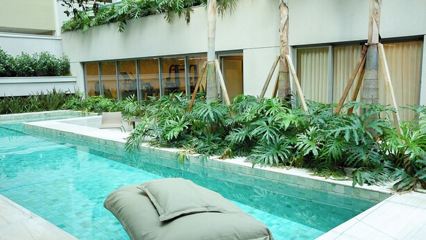 Ein Swimmingpool mit Liegestuhl und Kissen, umgeben von üppiger Bepflanzung und einem modernen Gebäude im Hintergrund.