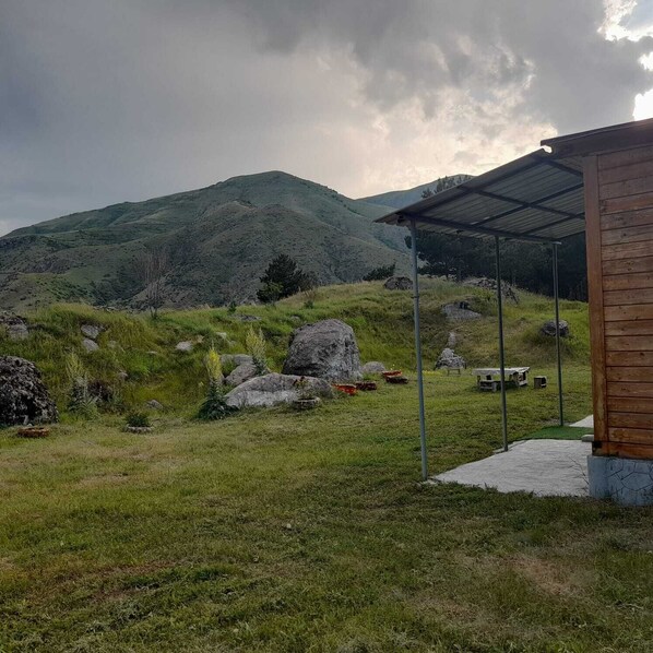 A rustic cabin with a metal roof and a wooden exterior, surrounded by a grassy landscape with large rocks and a hill in the background.