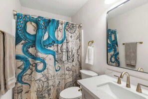 Une salle de bain avec un rideau de douche à motifs nautiques, des toilettes blanches, un lavabo avec des robinets dorés et un miroir qui reflète le rideau de douche.