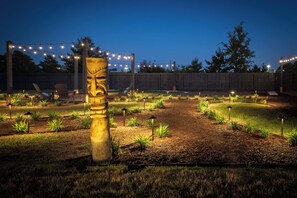 A landscaped backyard with a tiki head sculpture, string lights, and a wooden fence.