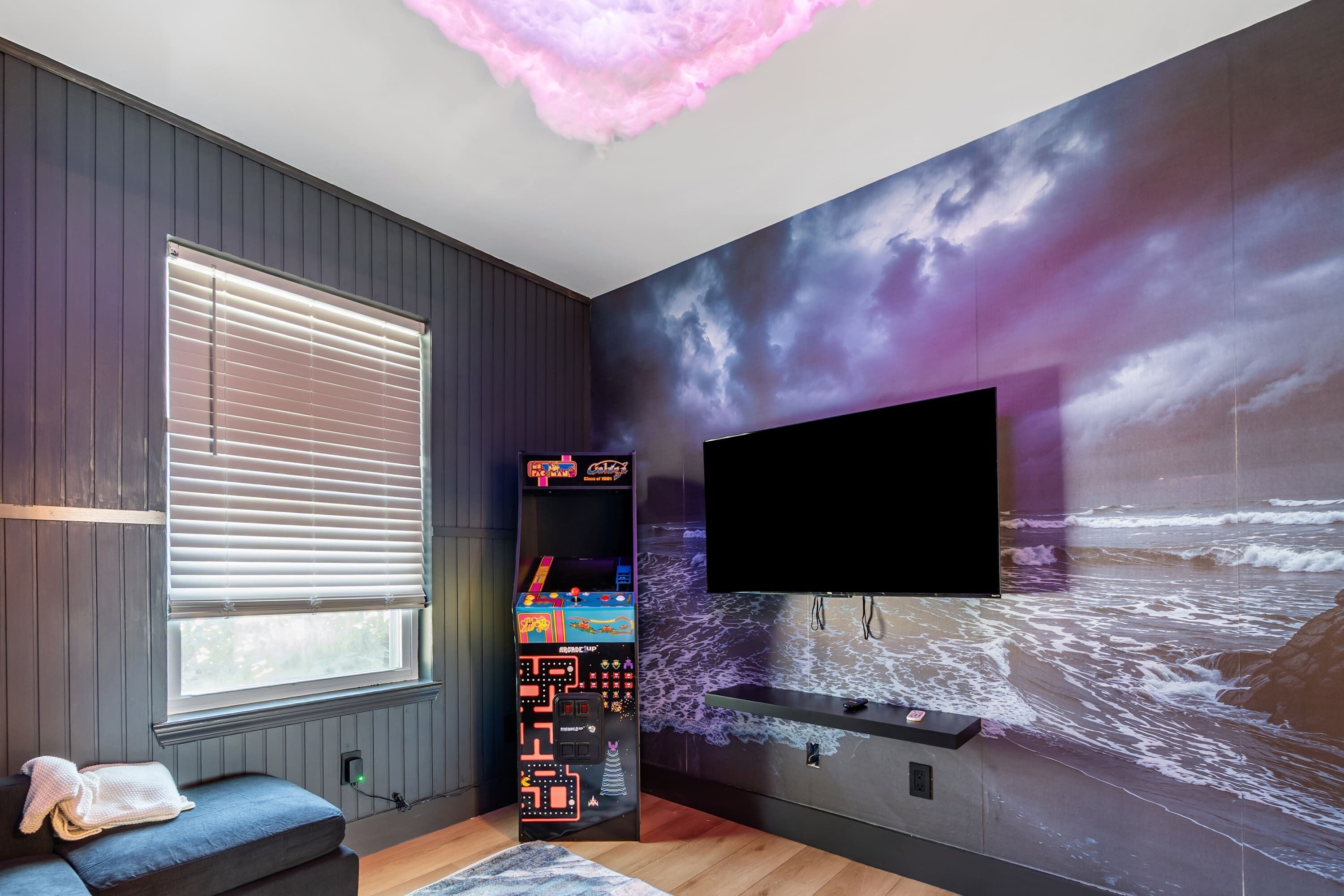 Ein Zimmer mit großem Fenster, Wand-Fernseher, Spielkonsole und Wandgemälde einer Strandansicht.