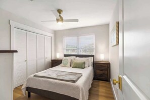 Une chambre à coucher comprenant un lit, des tables de chevet, un ventilateur de plafond, une fenêtre avec des stores et une porte avec une poignée.