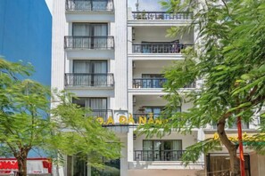Un edificio de varios pisos con balcones, un letrero que dice 'SA DANAM' y árboles en primer plano.