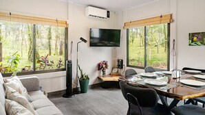 Ein modernes Wohnzimmer mit einer Couch, einem Esstisch für vier Personen, einem an der Wand montierten Fernseher und großen Fenstern mit Blick auf Grünflächen.