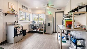 Eine kompakte Küche mit Gaskochfeld, Kühlschrank und Kaffeemaschine. Regale mit Geschirr und ein Fenster sorgen für natürliches Licht.