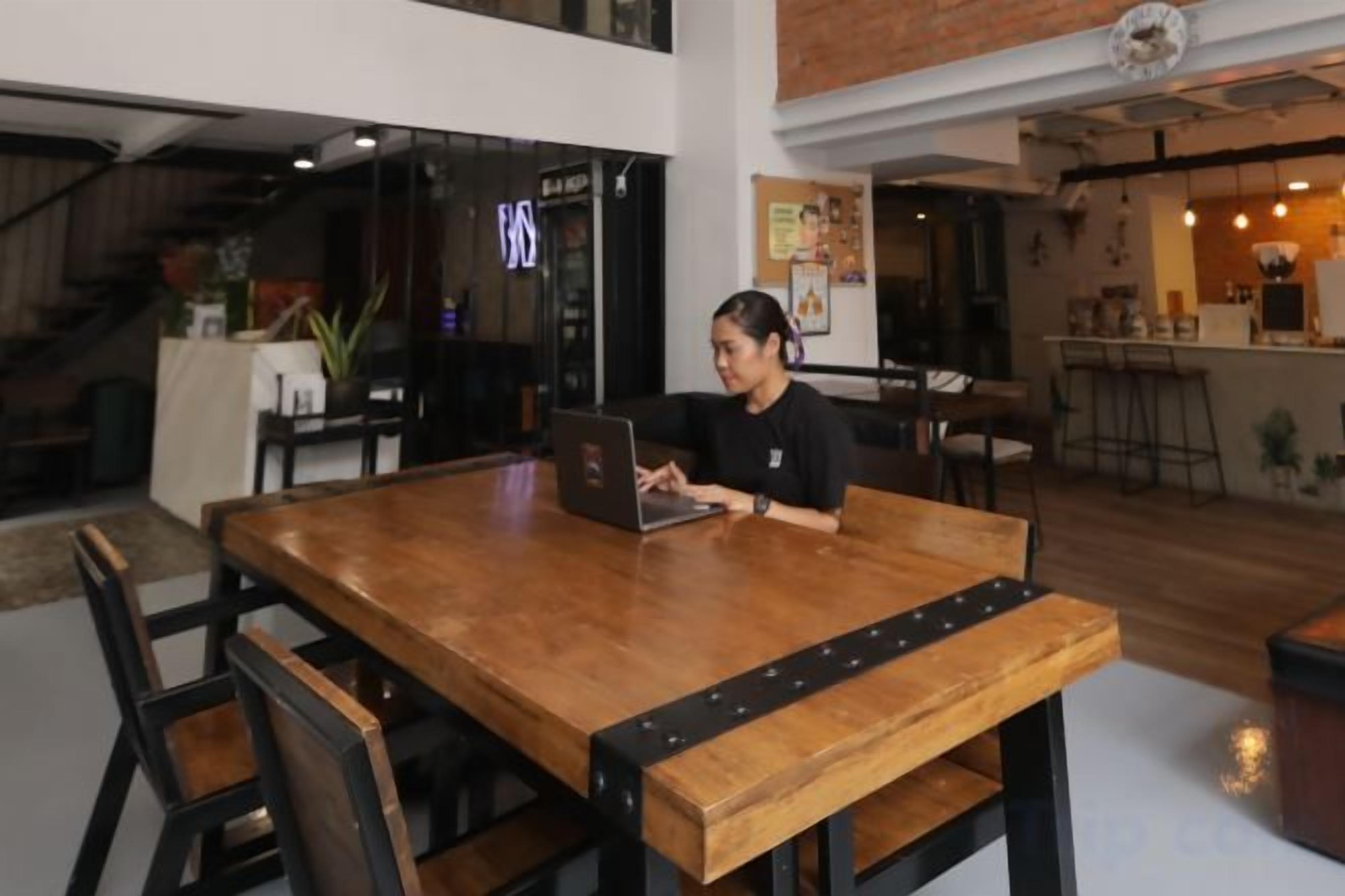 Une personne est assise à une table en bois, devant un ordinateur portable, dans une pièce qui comprend un bar et un escalier en arrière-plan.