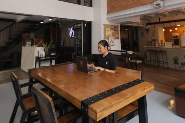 A person seated at a wooden table with a laptop, in a room with a bar area and a staircase in the background. - Wire Bangkok Prakhanong (Bangkok)