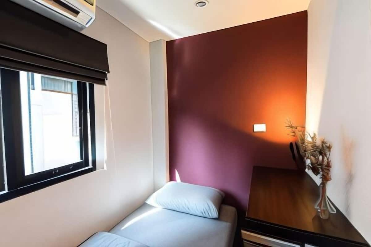 Een kleine eenpersoonskamer met een raam, een wandairconditioner en een nachtkastje met een vaas gedroogde bloemen.