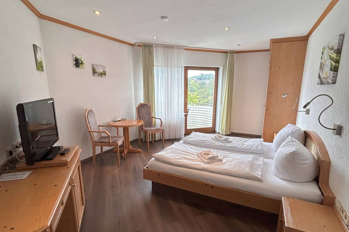 A hotel room with a bed, a desk with a television, a chair, and a view of a green landscape through the window.