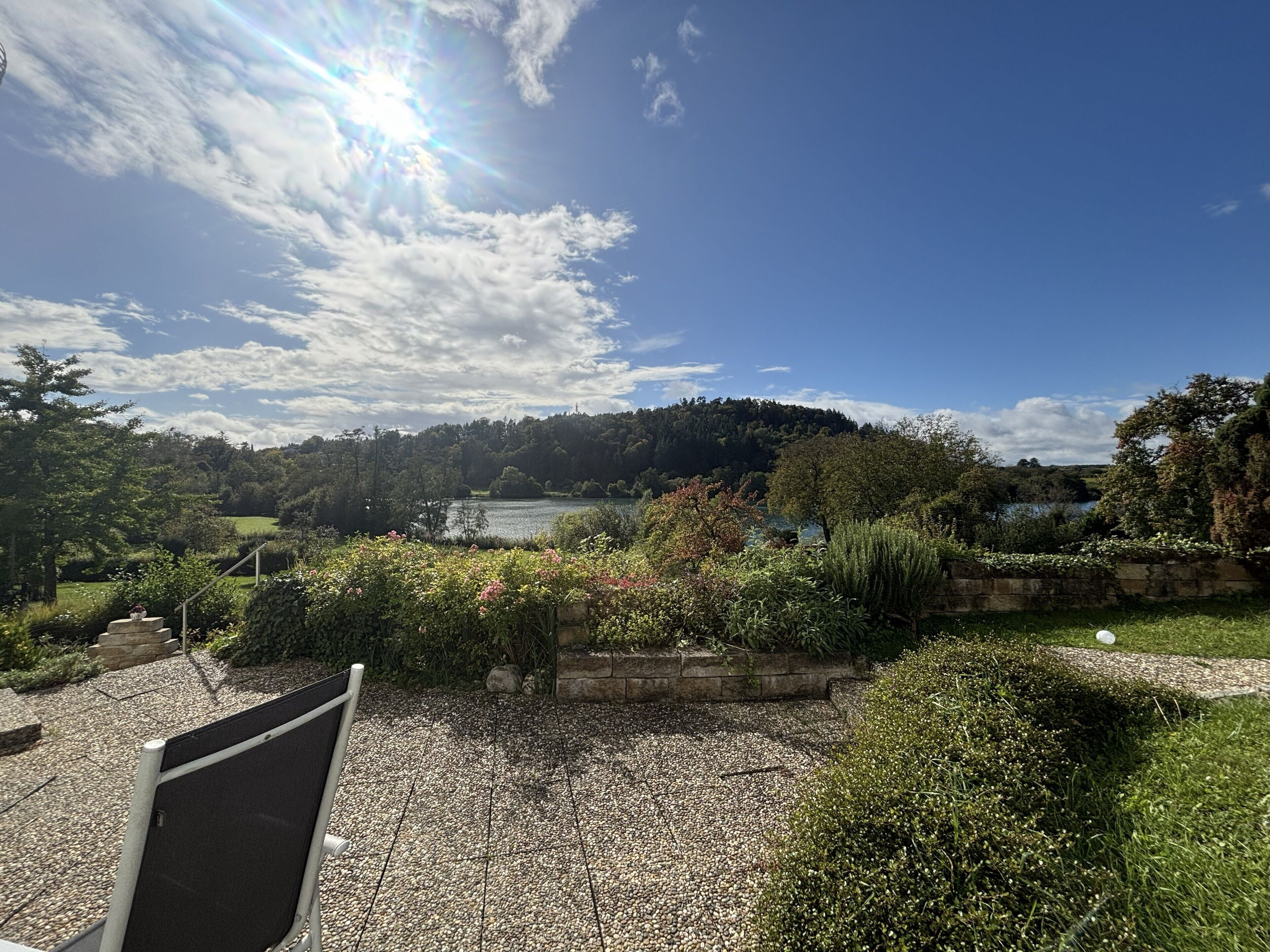 A garden with a gravel path, a stone wall, and a view of a lake and forested hill.