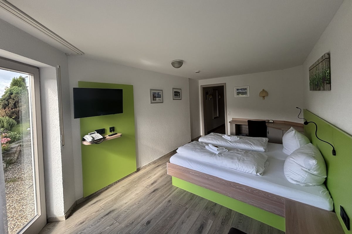 A modern hotel room with a bed, a TV mounted on a green wall, a small desk, and a bathroom visible through an open door.
