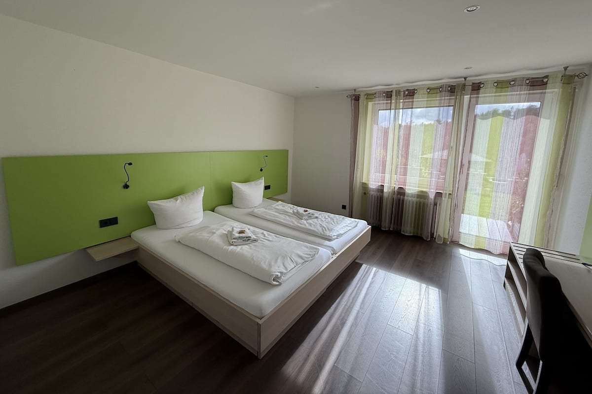 A hotel room with two beds, a green headboard, white bedding, and a large window with sheer curtains.