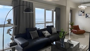 Ein modernes Wohnzimmer mit einer schwarzen Couch, einem Glastisch und Meerblick durch große Fenster.