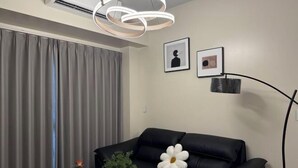 Ein modernes Wohnzimmer mit einem schwarzen Ledersofa, einem weißen Blütenkissen und einer kleinen Pflanze. An der Wand hängen zwei gerahmte Bilder, und es steht eine Stehlampe.