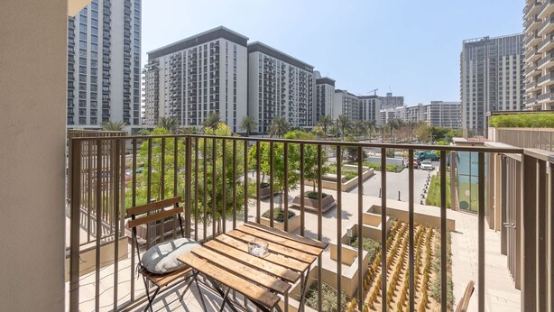 A balcony with a wooden table and bench, surrounded by a metal railing. In the background, there are high-rise buildings and a landscaped area with trees and a pool. - Dubai Hills Sleeps 4 With Awesome Swimming Pool (Dubai)
