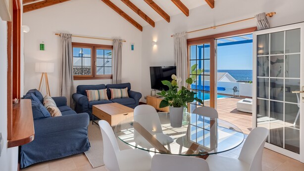 A living room with a sofa, coffee table, and TV, a glass table with chairs, and a sliding glass door to a balcony with a pool.