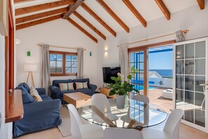A living room with a sofa, coffee table, and TV, a glass table with chairs, and a sliding glass door to a balcony with a pool. - Vacation Villa 'Turqué' with Sea View, Private Pool and Wi-Fi (Arrieta)