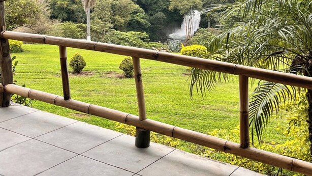 Vista desde un balcón techado con baranda de bambú, sobre un paisaje verde y exuberante con cascada y bosque denso.