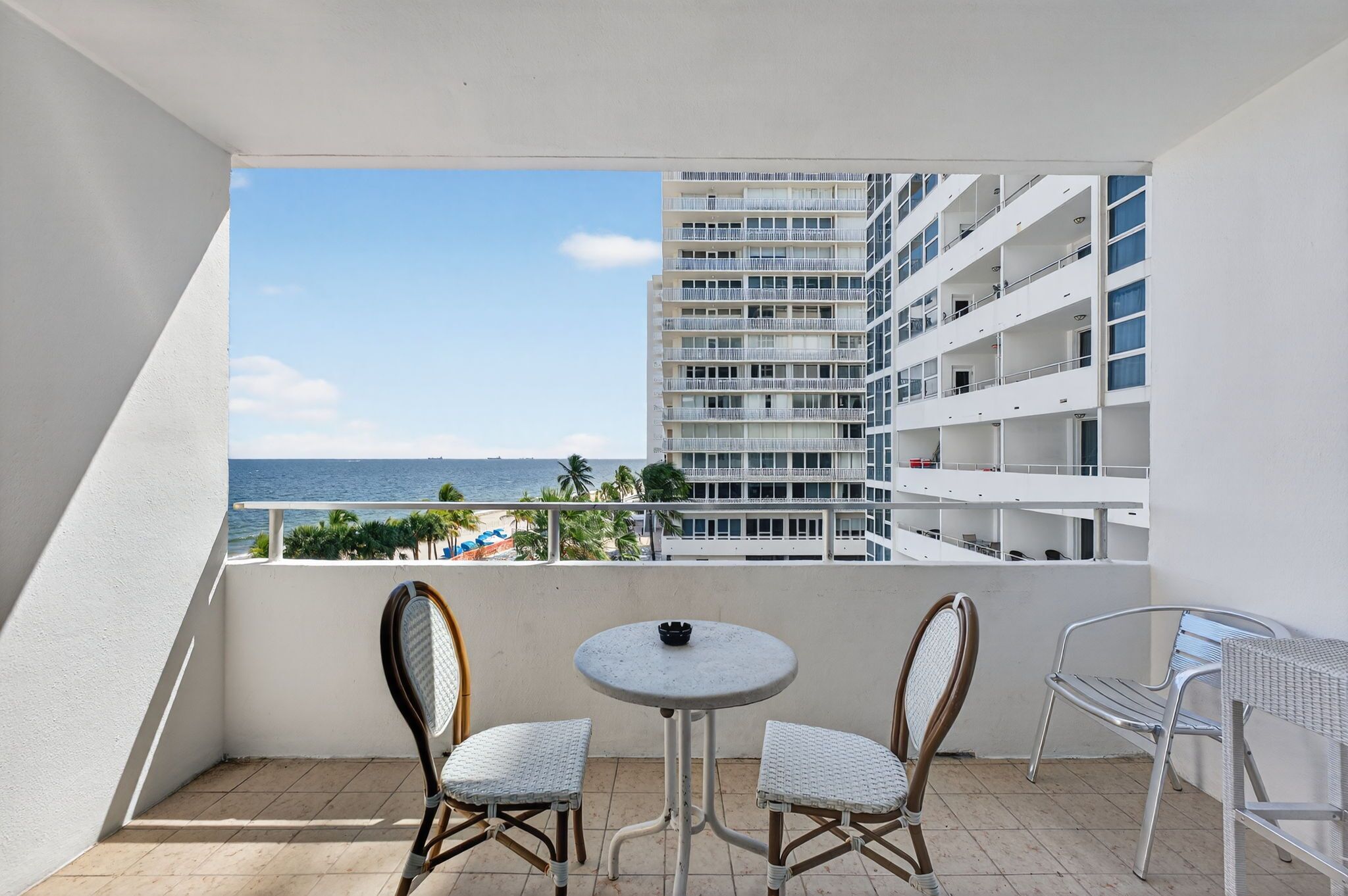 Een balkon met twee rieten stoelen en een klein rond tafeltje, met uitzicht op een strand en een hoog gebouw.